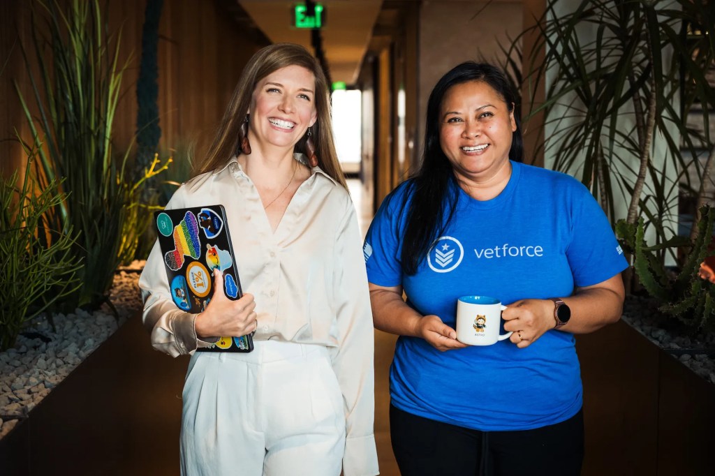 モダンなオフィスで立つ2人の女性。1人はコンピューターを操作し、1人はコーヒーマグを持っている