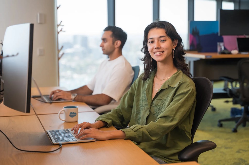 モダンなオフィスでデスクに座り、カメラに向かって微笑んでいる緑色のブラウスを着た女性