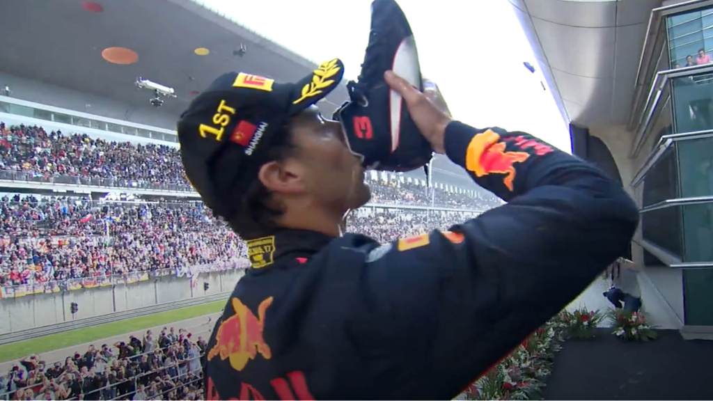F1 driver in Red Bull gear doing a shoey on the podium above a crowd.