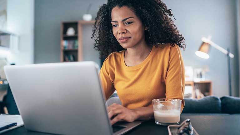 Une femme souriante avec des cheveux bouclés, portant un pull jaune, travaille sur son ordinateur portable tout en étant assise à un bureau avec un verre de lait à côté d'elle.
